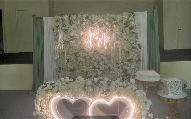 Wedding sweetheart table with a white flower wall, a neon 'You + Me' sign, and glowing heart lights