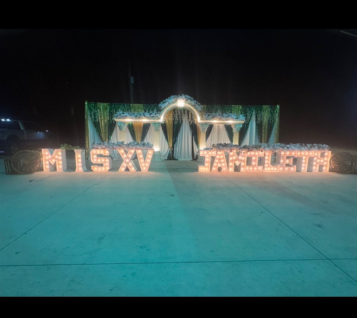 Outdoor quinceañera at night with 'MIS XV JAMILETH' light-up marquee letters and a green, gold, and white draped entrance